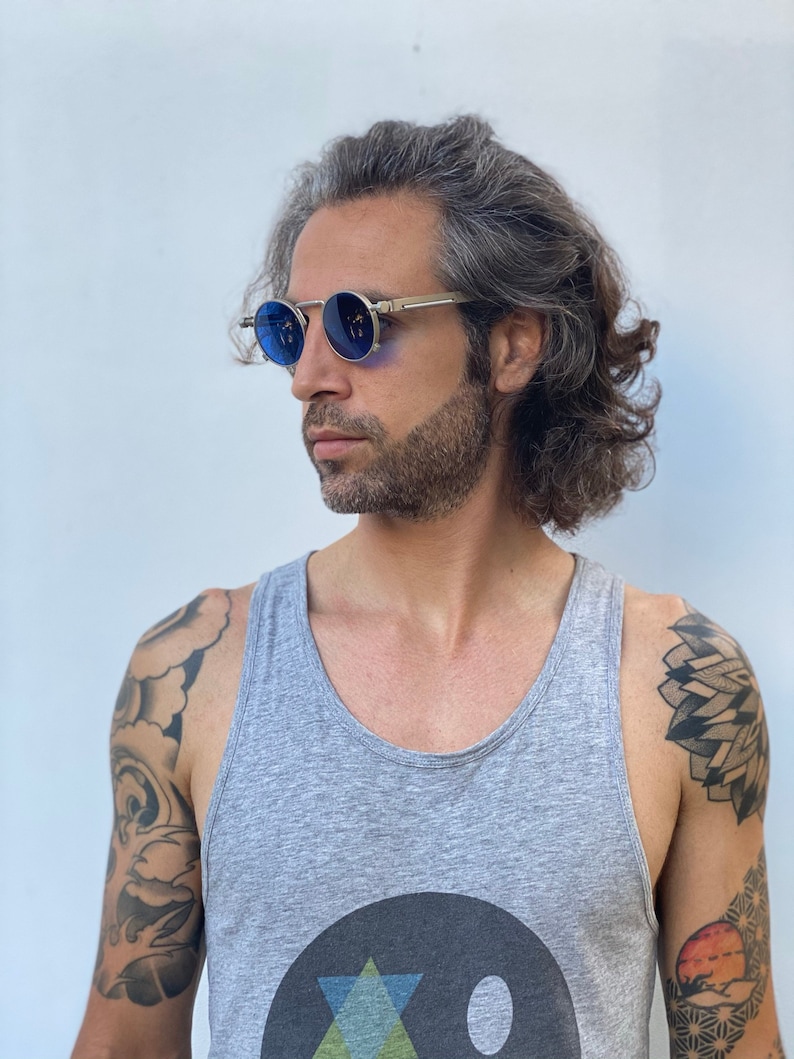 May include: A man with long gray hair wearing round blue sunglasses with a silver frame. He is wearing a gray tank top with a black circle and a geometric design on the front. He has tattoos on both arms.