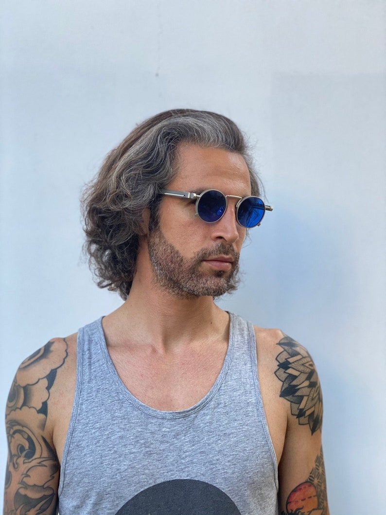 May include: A man with gray hair and a beard is wearing a gray tank top and blue round sunglasses. He has tattoos on his arms.