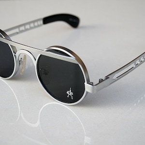Puede incluir: Gafas de sol redondas plateadas con lentes negros y un marco de metal plateado. Las gafas tienen un diseño único con una barra de metal en la parte superior de las lentes.
