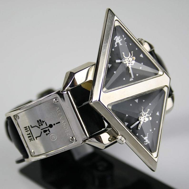 Pu&ograve; includere: Un orologio da polso triangolare argento e nero con un cinturino in pelle nera. Il quadrante dell'orologio ha un design unico con due finestre triangolari e il testo "ALEXANDER HI-TEIC" sul cinturino.