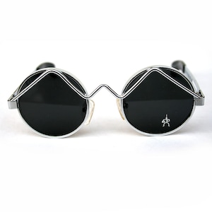 May include: Silver framed round sunglasses with black lenses. The sunglasses have a unique, angular design with a small symbol on the right lens.