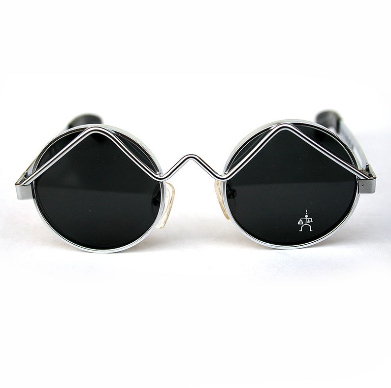 May include: Silver framed round sunglasses with black lenses. The sunglasses have a unique, angular design with a small symbol on the right lens.