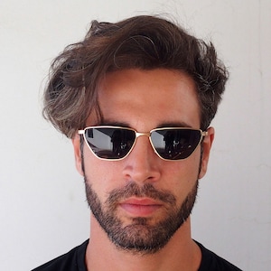 Peut inclure: Un homme aux cheveux bruns portant des lunettes de soleil à monture dorée avec des verres foncés. Les lunettes ont une forme œil de chat.