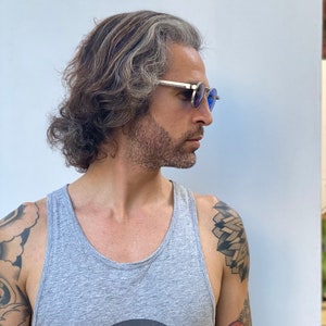 May include: A man with long, gray hair and a beard is wearing round, blue-tinted sunglasses. He is wearing a gray tank top with a black and white graphic design. He has tattoos on his arms.
