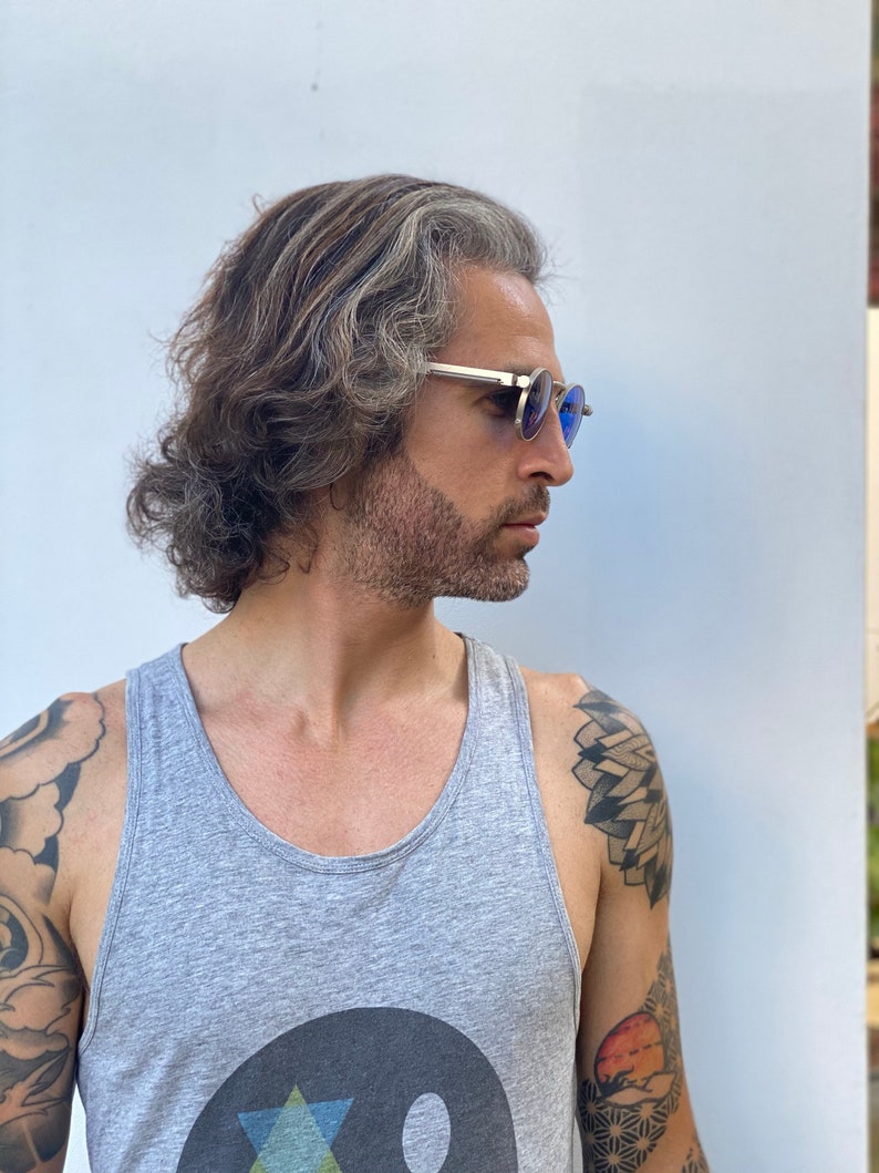May include: A man with long, gray hair and a beard is wearing round, blue-tinted sunglasses. He is wearing a gray tank top with a black and white graphic design. He has tattoos on his arms.