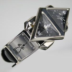 Pu&ograve; includere: Un orologio da polso argento e nero con un quadrante triangolare e un cinturino in pelle nera. Il quadrante dell'orologio ha un design argentato e il cinturino ha il testo "ALEXANDER HI-TEK" e un simbolo di marchio registrato.