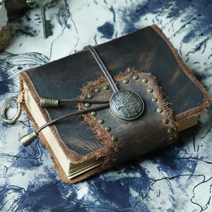 May include: A brown leather journal with a tree design on the front cover. The journal has a frayed edge and is decorated with metal studs and a leather strap with a metal clasp.
