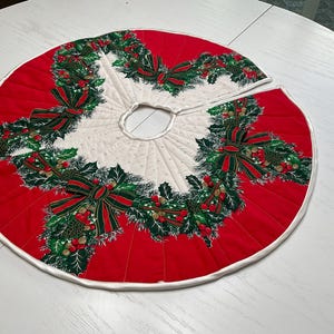 May include: A circular quilted Christmas tree skirt featuring a vibrant red outer border and a white center. The design includes a repeating pattern of green holly leaves, pine branches, red berries, and dark green bows with red trim.