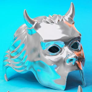Op de afbeelding: Een glanzend zilveren masker met een gehoornd ontwerp, met oogopeningen en een neus. Het masker heeft een reflecterend oppervlak en staat tegen een helderblauwe achtergrond. Het masker lijkt van metaal te zijn.