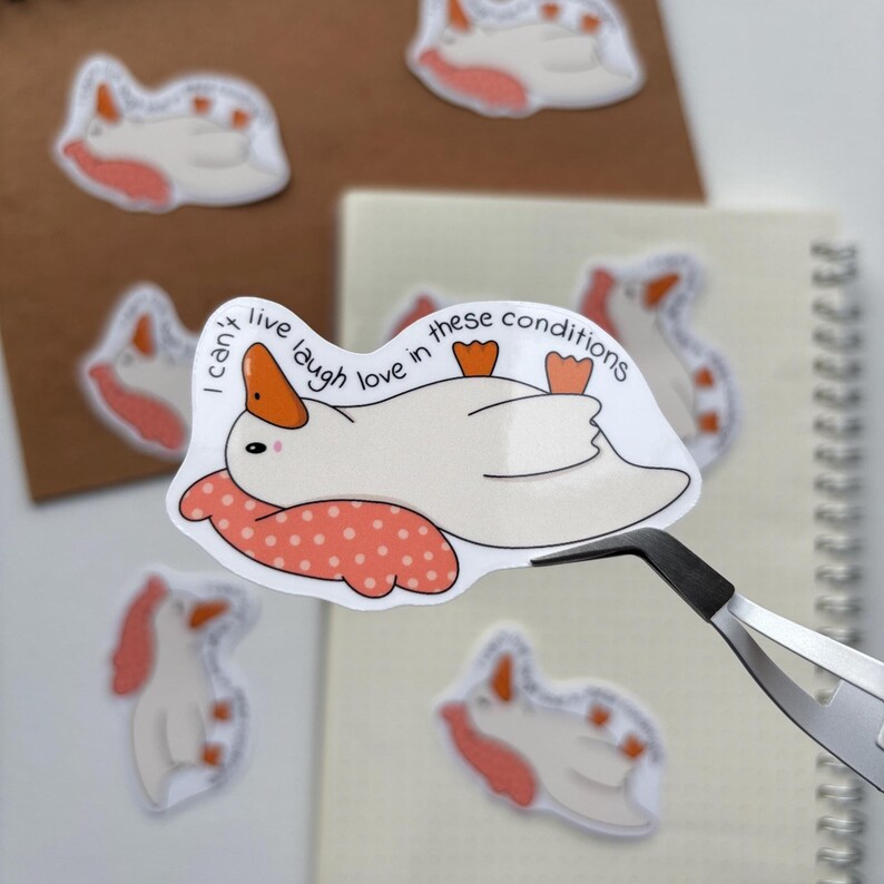 May include: A white sticker featuring a cartoon goose with an orange beak and feet, resting on a pink polka-dot pillow. The text "I can't live laugh love in these conditions" is written above the goose. Several similar stickers are visible.
