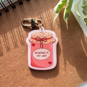 Peut inclure: Un porte-clés en acrylique rose en forme de pot de confiture, avec un fermoir doré. Le pot a un couvercle avec des détails de cœur et un nœud rouge. Le texte "READING IS MY JAM" est imprimé sur le devant, avec deux cœurs rouges.