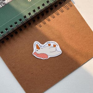 May include: A white sticker featuring a cartoon goose lying down on a pink pillow with white polka dots. The sticker has the text "I can't live laugh love in these conditions" in a circular pattern around the goose.