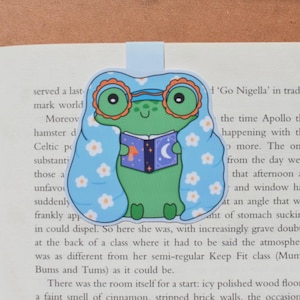 Könnte beinhalten: Ein skurriles Lesezeichen mit einem grünen Frosch mit Brille und einer blauen Decke mit weißen Blumen. Der Frosch liest ein Buch mit Mond- und Pilzdesign. Das Lesezeichen ist aus Papier.