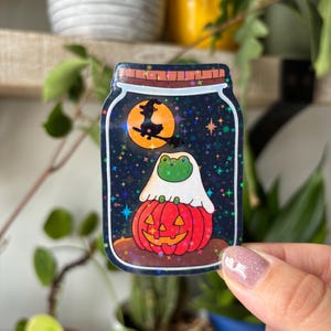 Może przedstawiać: Naklejka o tematyce Halloween przedstawiająca żabę w kostiumie ducha siedzącą na wyrzeźbionej dyni w słoiku. Słoik ma tło gwiaździstego nieba z wiedźmą lecącą obok pełni księżyca. Naklejka ma efekt holograficzny.