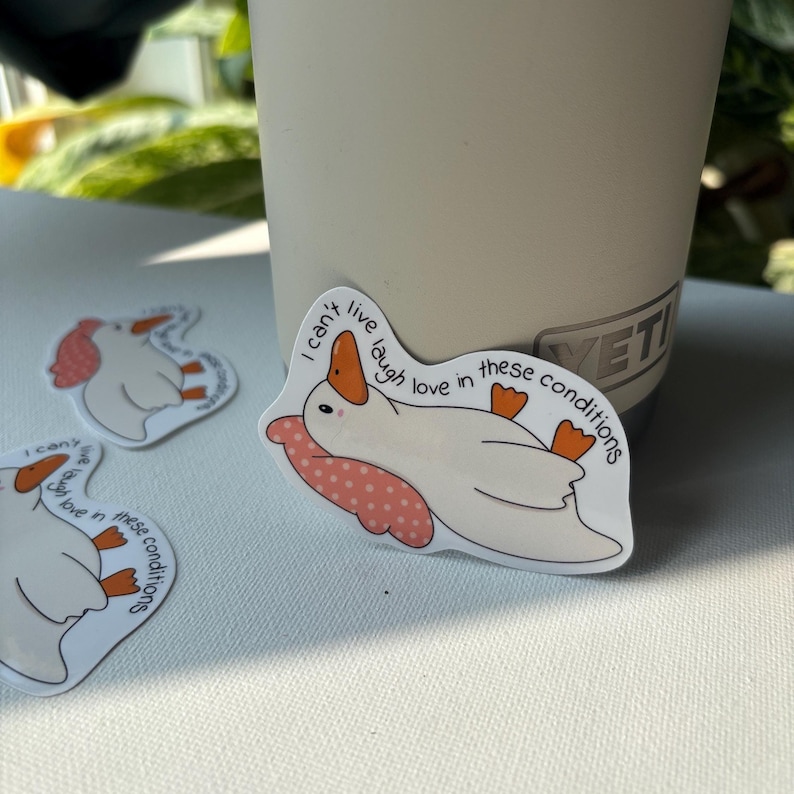 May include: White duck sticker with orange beak and feet, resting on a pink polka-dot pillow. The sticker has the text "I can't live laugh love in these conditions". Several stickers are visible.