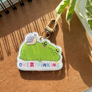 Peut inclure: Un porte-clés en acrylique transparent représentant une grenouille verte avec un escargot sur le dos. La grenouille a des joues roses et le mot "OVERTHINKING" en lettres colorées. Un fermoir doré est attaché.