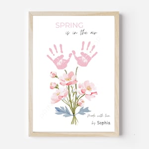 Könnte beinhalten: Ein gerahmter Druck in einem hellen Holzrahmen. Das Kunstwerk zeigt rosa Handabdrücke über einem Strauß rosa Blumen mit grünen Stielen und blauen Blättern. Der Text "SPRING is in the air" und "Made with love, by Sophia" sind ebenfalls enthalten.