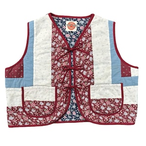May include: A quilted vest featuring a patchwork design with floral patterns in red, white, and blue. The garment has a V-neckline, three front tie closures, and two patch pockets. The interior lining is dark blue with a small floral print.