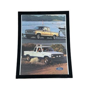 May include: Framed vintage print featuring two Ford pickup trucks. The top truck is gold, parked near water with a boat. The bottom truck is white with blue accents, driving on a dirt road. The Ford logo is visible.