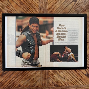 May include: A framed magazine article featuring a portrait of a person wearing a black studded leather vest and matching wristband, alongside a smaller photograph of two boxers in a ring. The text includes the headline Now Here's A Macho, Macho, Macho Man.