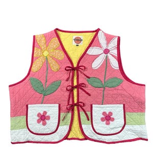 May include: A quilted vest in pink with a yellow interior lining, featuring three front tie closures. The vest displays large appliquéd flowers in orange and white with green leaves, and two white pockets each decorated with a small pink flower.