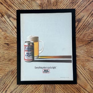 May include: A framed vintage advertisement featuring a silver Bud Light beer can next to a glass mug filled with golden beer and white foam. The text reads Everything else is just a light. with the Bud Light logo centered at the bottom.