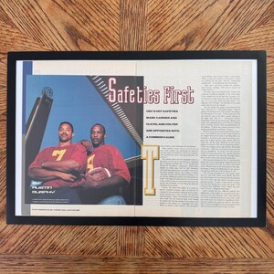 Puede incluir: Un artículo de revista enmarcado titulado Safeties First que presenta una fotografía de dos jugadores de fútbol americano con camisetas rojas, uno de ellos sosteniendo un balón. El texto incluye el titular USC'S HOT SAFETIES MARK CARRIER AND CLEVELAND COLTER ARE OPPOSITES WITH A COMMON CAUSE.