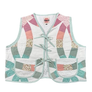 May include: A sleeveless quilted vest featuring a white base with a patchwork pattern of pink, sage green, and floral fabric segments. The vest has a light green trim, three front tie closures, and two patch pockets on the lower front panels.