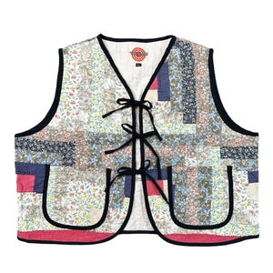 May include: A sleeveless vest featuring a patchwork design of various floral and geometric patterns in shades of pink, blue, white, and brown. The vest has a V-neckline, three black tie closures, two front pockets, and black trim. A logo is on the inner neck.