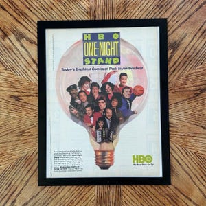Op de afbeelding: Een ingelijste vintage advertentie voor HBO One-Night Stand met een gloeilampafbeelding gevuld met een groep komieken. De tekst luidt HBO One-Night Stand, Today's Brightest Comics at Their Inventive Best en HBO The Best Time On TV.