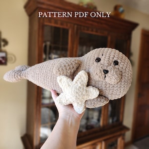 May include: A crocheted plush seal toy holding a crocheted starfish. The seal is brown and the starfish is cream colored. The toy is being held by a hand.