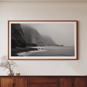 May include: A black and white framed print of a coastal landscape with cliffs and ocean. The print is displayed above a mid-century modern wooden sideboard with three drawers and two cabinet doors. A vase with decorative branches sits on the sideboard.