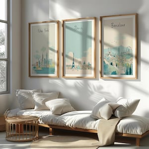 May include: Three framed watercolor prints depicting cityscapes of New York, Paris, and Barcelona. Each print features a light-colored background with a city skyline and the city's name in a handwritten font. The frames are made of light-colored wood.