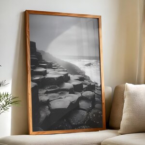 May include: Black and white framed print of a coastal landscape. The image features a series of hexagonal rock formations leading to the ocean. The frame is made of light brown wood. The print is leaning against a wall.