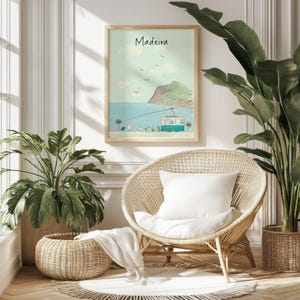 May include: A framed print with the word "Madeira" in a script font. The artwork depicts a coastal scene with a teal cable car, blue water, and a green mountain. The print is displayed above a wicker chair with a white cushion, with plants in the foreground.