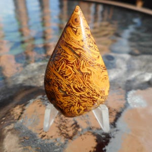 May include: A teardrop-shaped gemstone with a polished surface. The stone features a swirling pattern of golden yellow and reddish-brown hues. It is displayed on a clear, acrylic stand, showcasing its unique natural design. The gemstone is likely used for jewelry or decorative purposes.
