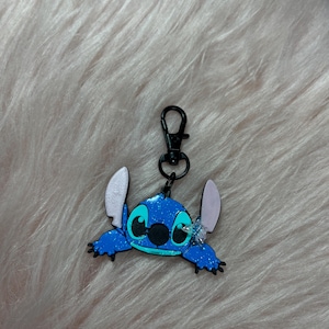 May include: A blue Stitch keychain with a black clasp. The keychain features a glittery blue Stitch character with large ears and a black nose. The background is a soft, pinkish-white fur.