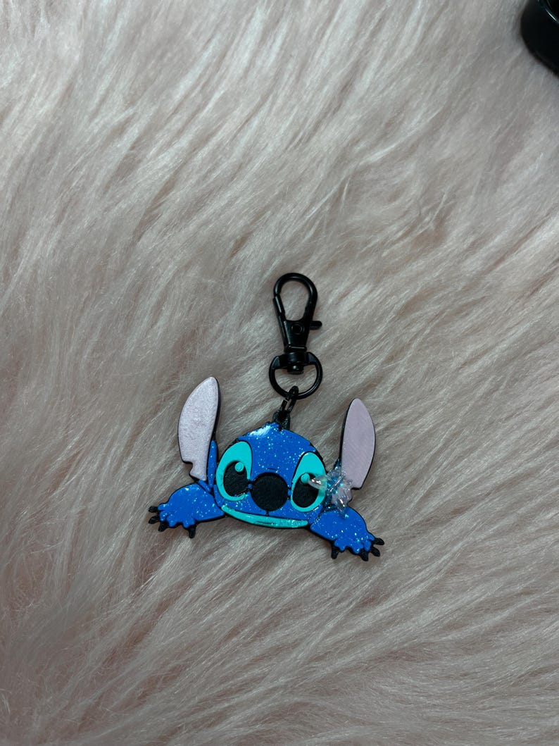May include: A blue Stitch keychain with a black clasp. The keychain features a glittery blue Stitch character with large ears and a black nose. The background is a soft, pinkish-white fur.