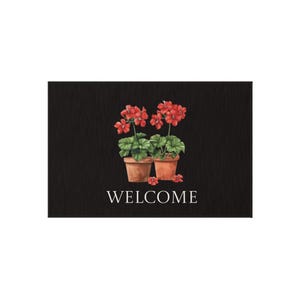 Op de afbeelding: Een zwarte deurmat met een aquarelontwerp met twee geraniums in pot met rode bloemen en groene bladeren. Het woord "WELCOME" staat eronder in het wit gedrukt.