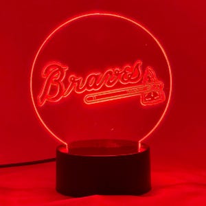May include: A red LED lamp featuring the word "Braves" in a script font, along with a tomahawk logo. The lamp has a circular design and sits on a black base. The background is also red.