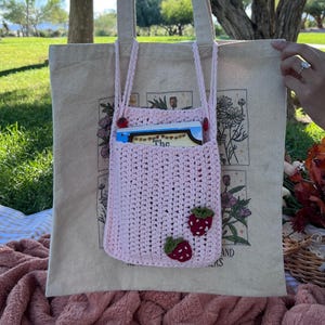 May include: A light pink crochet tote bag with a matching pocket and strawberry appliqués. The tote bag is inside a beige canvas bag with a floral design. A book is visible in the pocket. The bag has beige straps.