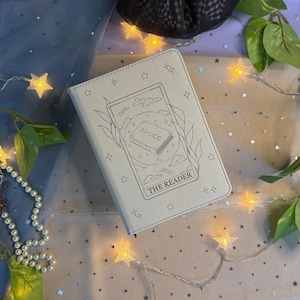 Op de afbeelding: Een lichtgrijs notitieboek met een zwart-wit illustratie van een boek en de tekst "THE READER". Het notitieboek is omgeven door lichtslingers en groen, met een zwarte scrunchie op de achtergrond.