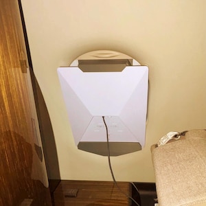 May include: A white, geometric-shaped device with a black cable, mounted on a light-colored wall. The device has a multi-faceted design and is positioned above a wooden surface. A tan seat is visible on the right.