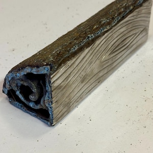 May include: A rectangular, metallic sculpture with a textured, wood-grain pattern on one side. The top edge has a rough, dark brown and blue-tinged surface. The end reveals an intricate, swirling interior design. The overall color is silver.