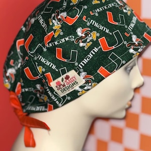 May include: A green scrub cap featuring the Miami Hurricanes logo and text. The cap has a repeating pattern of the team's logo and the word "Miami" in orange and white. The cap is tied with an orange ribbon. The tag reads "Sew Sassy Designs".