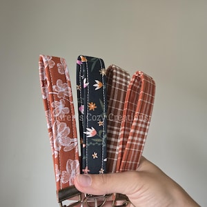 May include: Four fabric wristlet keychains featuring various patterns including a rust orange floral print, a black floral print, a brown and white plaid, and a rust orange and white gingham, each with a metal clasp and a circular key ring.