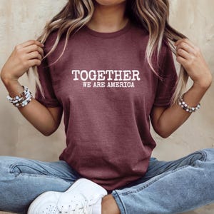 Może przedstawiać: Bordowa koszulka z napisem "TOGETHER WE ARE AMERICA" w białych literach. Koszulka ma okrągły dekolt i wydaje się być wykonana z miękkiego materiału. Osoba ma na sobie niebieskie dżinsy i białe trampki.