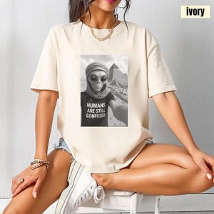 Op de afbeelding: Ivoor kleurig T-shirt met een zwart-wit afbeelding van een alien die een selfie maakt voor de piramides. Op het shirt van de alien staat "HUMANS ARE STILL CONFUSED". De top heeft korte mouwen en een ronde hals.