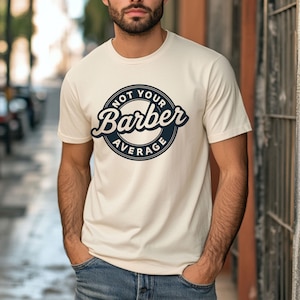 Pode incluir: Camiseta creme com o texto "NOT YOUR Barber AVERAGE" em um design circular azul marinho. A camiseta de manga curta tem gola redonda. A pessoa que usa a camiseta tem as mãos nos bolsos.