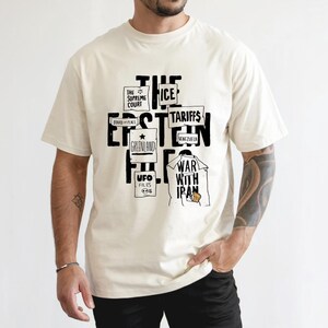 Puede incluir: Camiseta color crema con un diseño gráfico negro. El diseño incluye las palabras "THE EPSTEIN FILES" y otros elementos de texto como "ICE", "Tariffs", "War with Iran" y "UFO Files".
